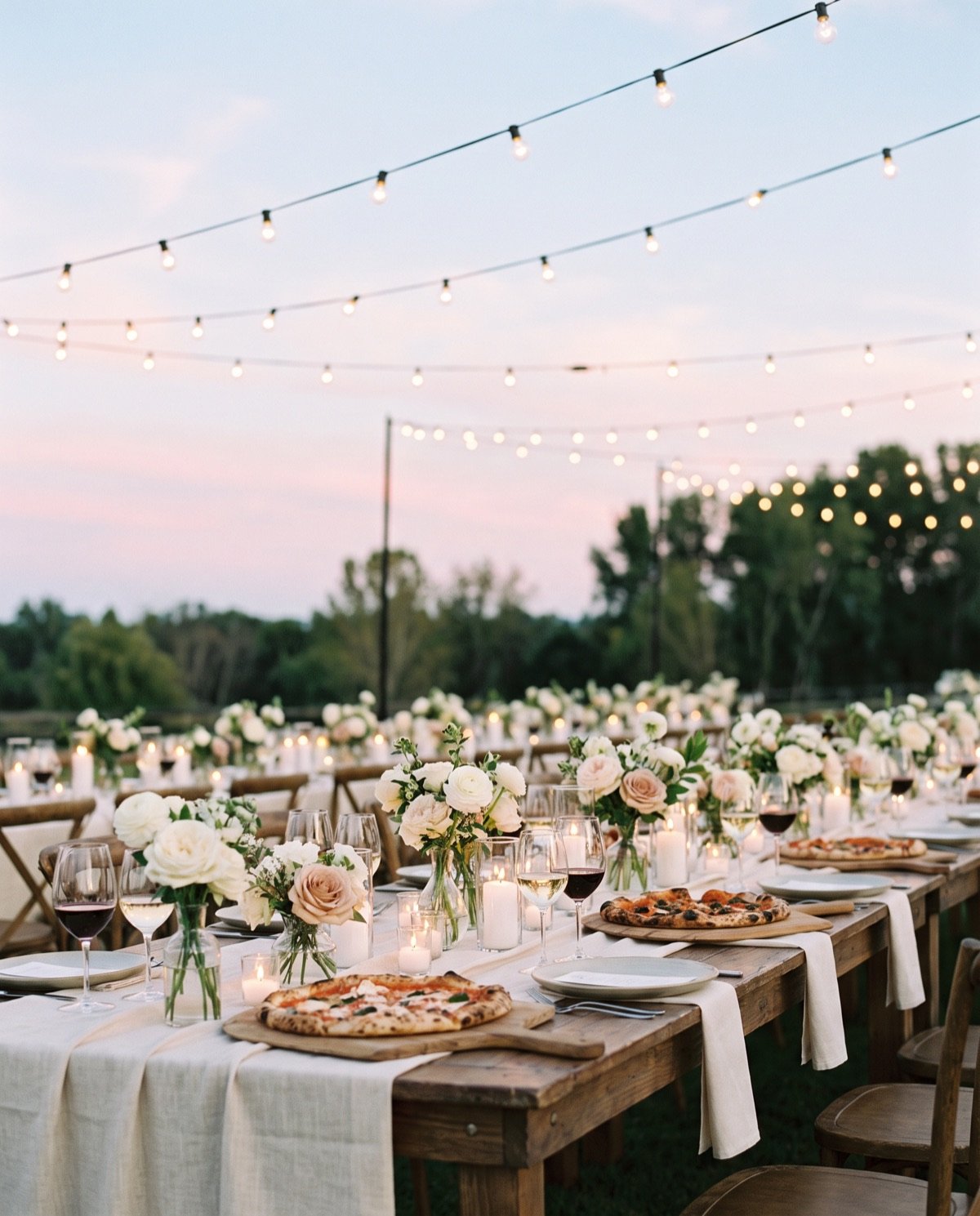 Long outdoor wedding table at dusk with cream linens, flowers, candles and wood-fired pizzas under string lights