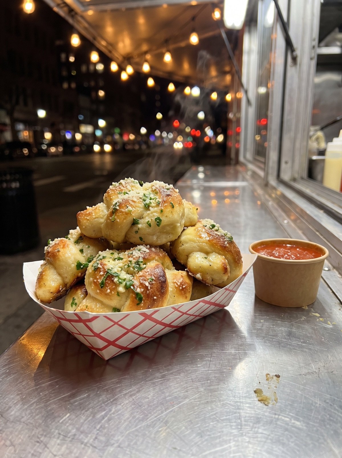 Garlic knots with parsley and parmesan in a checkered paper boat, with a cup of red sauce